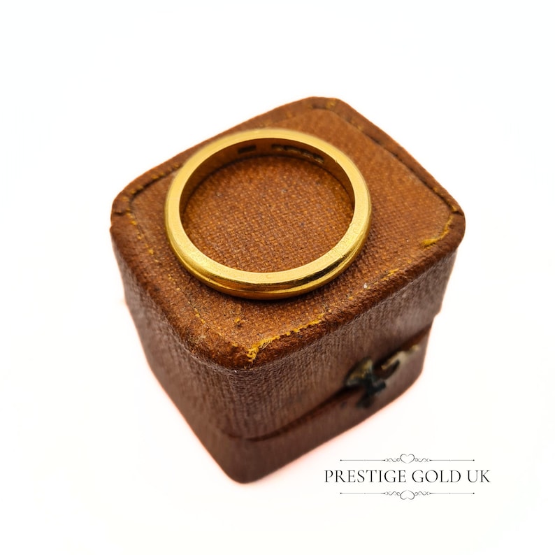 May include: A gold wedding band in a brown velvet ring box. The box is closed and the ring is visible on top.