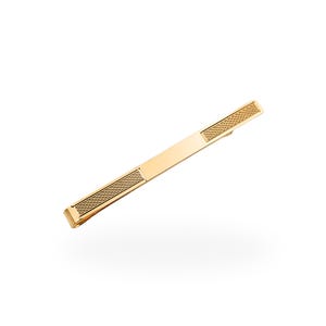 May include: A gold-toned metal tie bar featuring a smooth, polished center section flanked by two rectangular panels with a fine, cross-hatched textured pattern. The accessory has a sleek, elongated rectangular shape with a spring-loaded clip mechanism.