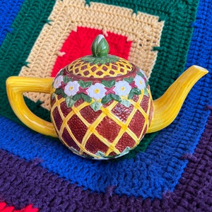 May include: A yellow ceramic teapot with a brown and yellow patterned design. The teapot has a lid with a green flower on top. The teapot is sitting on a colorful crocheted surface.
