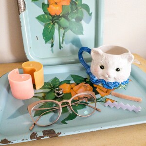 May include: A blue metal tray with a floral design, a white ceramic cat mug, a pair of gold-rimmed glasses, two candles, and a pink and purple hair clip.