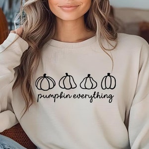 Peut inclure: Un sweat-shirt crème avec un dessin au trait noir de quatre citrouilles et l'inscription "pumpkin everything". Le sweat-shirt est de couleur neutre, parfait pour l'automne.