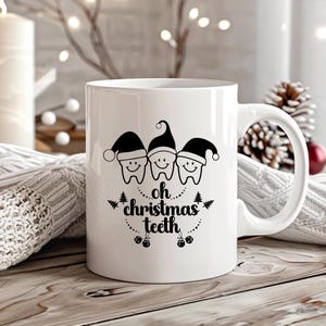 May include: White ceramic mug with a black graphic of three smiling teeth wearing Santa hats. The text "oh christmas teeth" is below the teeth. The mug is on a wooden surface with a blurred background of lights and a pinecone.