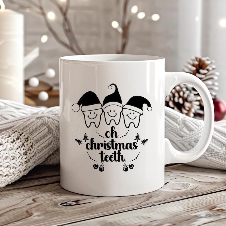 May include: White ceramic mug with a black graphic of three smiling teeth wearing Santa hats. The text "oh christmas teeth" is below the teeth. The mug is on a wooden surface with a blurred background of lights and a pinecone.