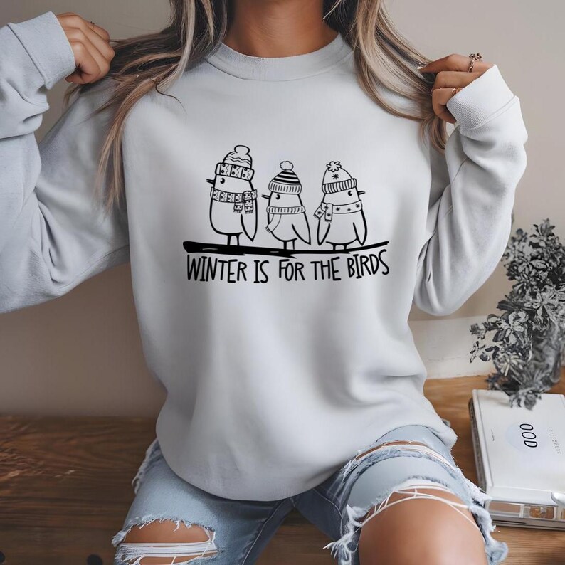 Op de afbeelding: Een grijs sweatshirt met een zwarte afbeelding van drie vogels die wintermutsen en sjaals dragen. De tekst "WINTER IS FOR THE BIRDS" is onder de vogels gedrukt.