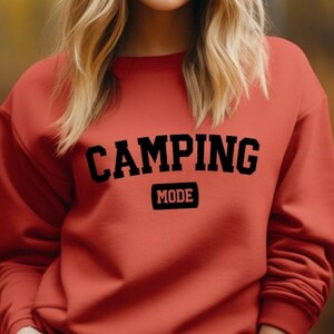 Op de afbeelding: Een terracotta-kleurige sweater met ronde hals waarop het woord CAMPING in grote zwarte blokletters over de borst is gebogen, met het woord MODE in een kleiner zwart lettertype in een rechthoekig kader direct onder de tekst.