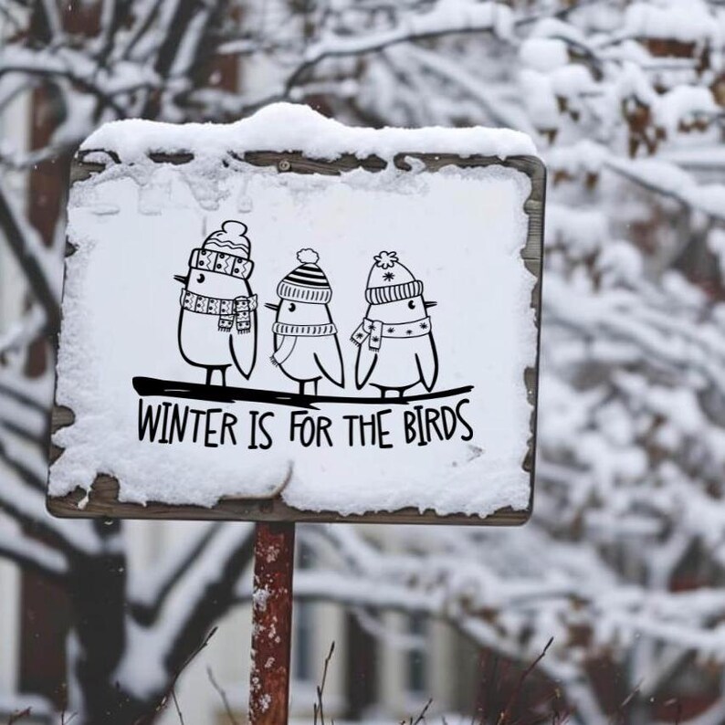 Op de afbeelding: Een wit bord met zwarte tekst en illustraties van drie vogels die hoeden dragen. Het bord staat "Winter is for the birds". Het bord is bedekt met sneeuw en is bevestigd aan een houten paal.