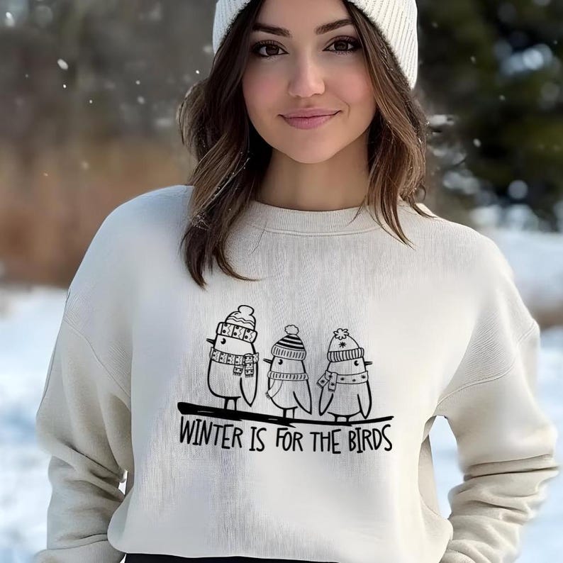 Op de afbeelding: Een wit sweatshirt met een zwarte afbeelding van drie vogels die wintermutsen en sjaals dragen. De tekst "Winter is for the birds" staat onder de afbeelding.