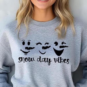 Op de afbeelding: Een lichtgrijs sweatshirt met een afbeelding van drie lachende gezichten met snorren. De tekst "snow day vibes" is in het zwart onder de gezichten gedrukt. De sweatshirt heeft een ronde hals.