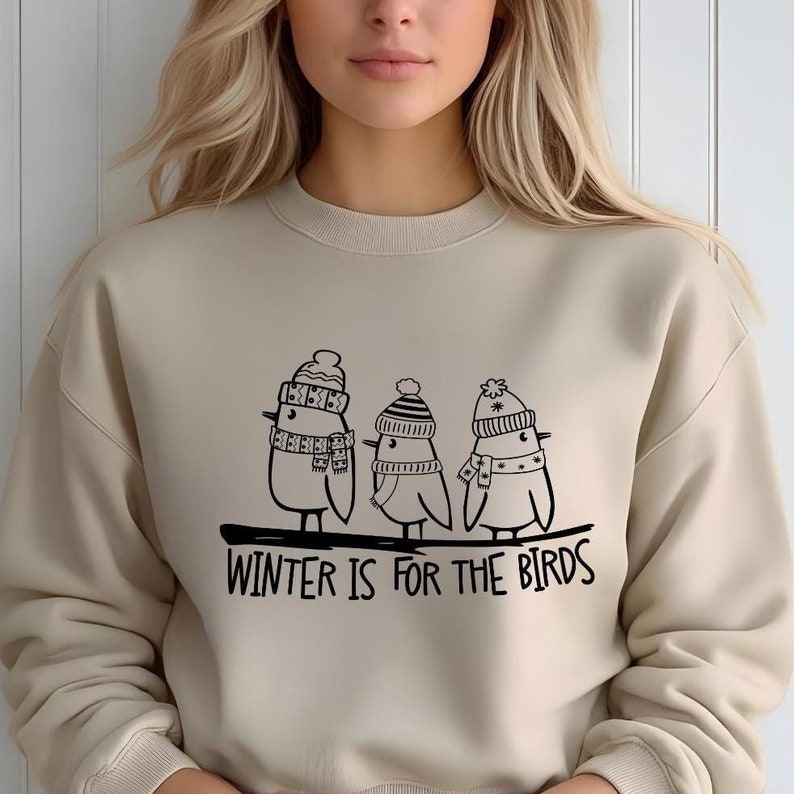Op de afbeelding: Een beige sweatshirt met een zwarte afbeelding van drie vogels die wintermutsen en sjaals dragen. De tekst "WINTER IS FOR THE BIRDS" is onder de vogels gedrukt.
