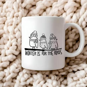 Op de afbeelding: Witte keramische mok met een zwart-witte illustratie van drie vogels die wintermutsen en sjaals dragen. De tekst "WINTER IS FOR THE BIRDS" is onder de vogels gedrukt.