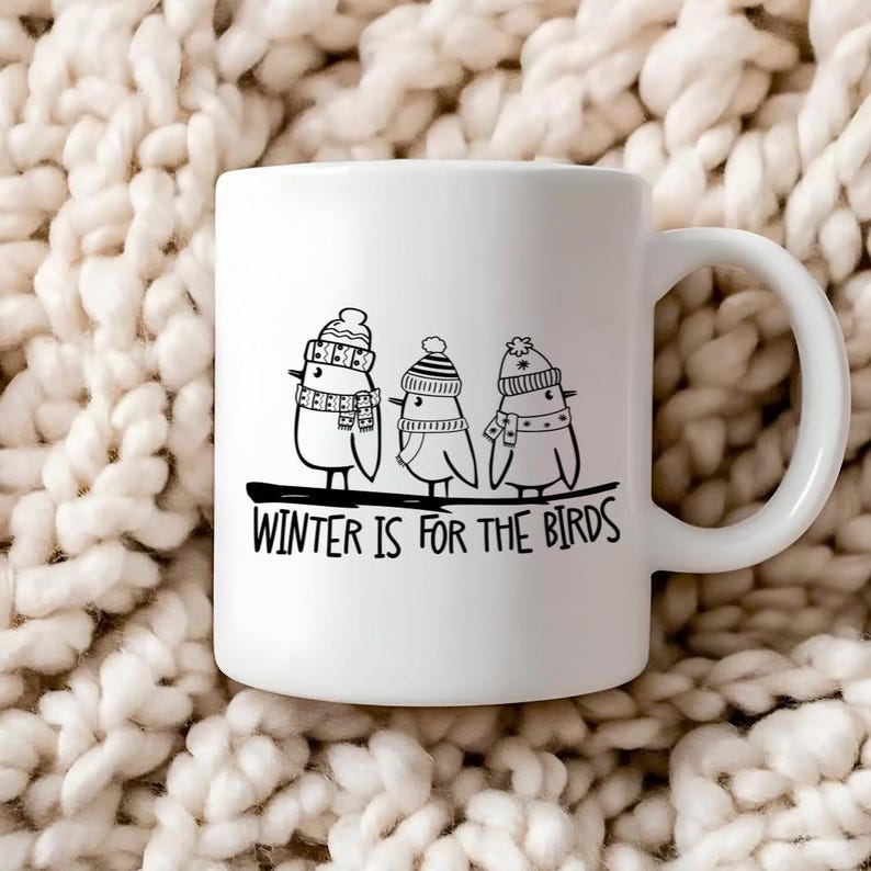 Op de afbeelding: Witte keramische mok met een zwart-witte illustratie van drie vogels die wintermutsen en sjaals dragen. De tekst "WINTER IS FOR THE BIRDS" is onder de vogels gedrukt.