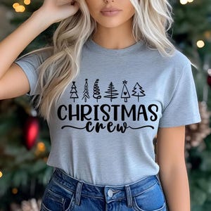 May include: A light gray t-shirt with the text "CHRISTMAS crew" in black script. Above the text are six stylized Christmas tree designs. The shirt is short-sleeved and made of a soft material.