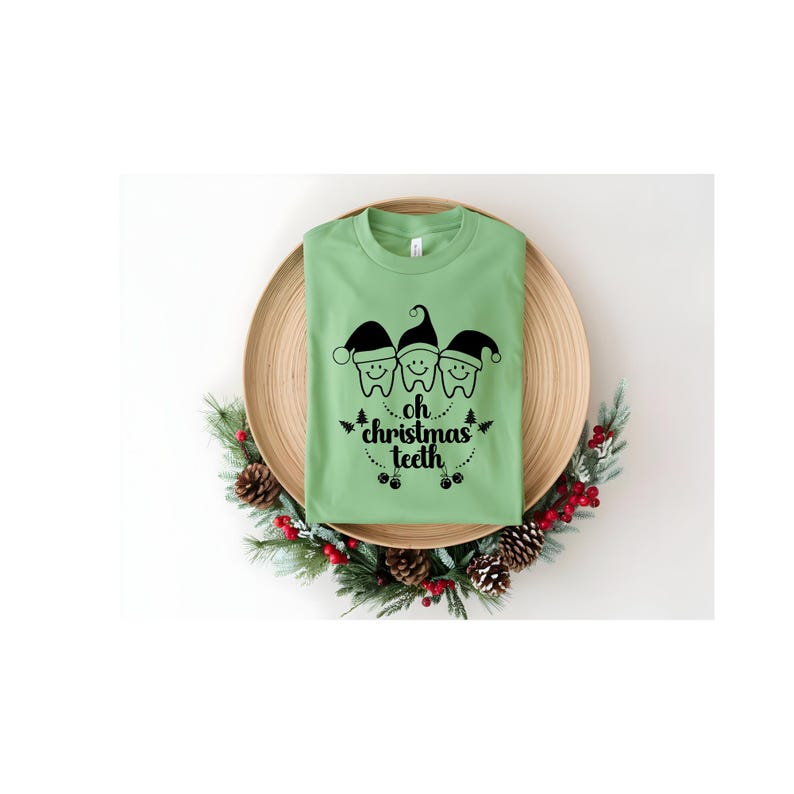 May include: A light green t-shirt with a black graphic of three teeth wearing Santa hats and the text "oh christmas teeth". The shirt is displayed on a wooden plate surrounded by a Christmas wreath.