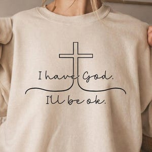 Op de afbeelding: Een beige sweater met ronde hals en een zwarte lijntekening van een christelijk kruis. De tekst "I have God." staat boven een gebogen lijn, met de tekst "I'll be ok." eronder gedrukt in een eenvoudig, cursief lettertype.