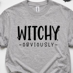 May include: A heather gray crew neck t-shirt featuring the black text WITCHY, OBVIOUSLY in a stylized font. The word WITCHY is flanked by two small four-pointed star icons, and the word OBVIOUSLY is centered below with a dash on each side.