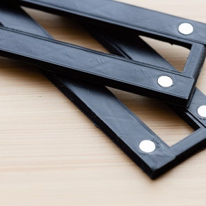 May include: Black, accordion-style, expanding ruler with white circular rivets. The ruler is made of a matte black material and is partially extended, resting on a light wood surface. The design suggests a tool for measuring or drafting.