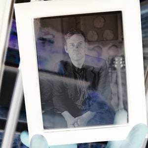 May include: A white framed photograph featuring a man in a dark blazer and striped shirt. The image is held by a gloved hand, with a blurred background including a guitar and circular objects.