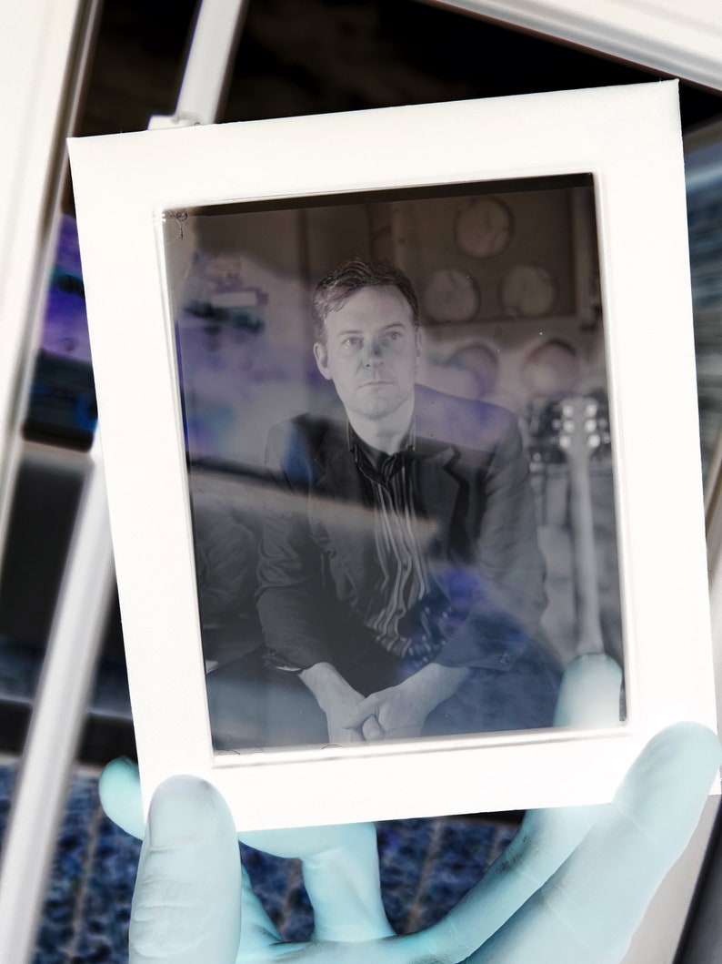 May include: A white framed photograph featuring a man in a dark blazer and striped shirt. The image is held by a gloved hand, with a blurred background including a guitar and circular objects.