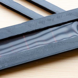 May include: Black rectangular frame with a textured surface and a clear window. The window has red lines. Additional black strips are visible. The items are on a light wood surface.