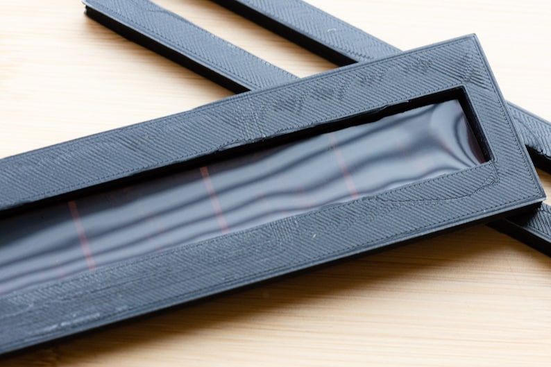May include: Black rectangular frame with a textured surface and a clear window. The window has red lines. Additional black strips are visible. The items are on a light wood surface.