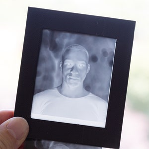 May include: A black and white photographic print of a person in a black frame. The image is a negative, showing a light-colored face and upper body against a darker background. The frame is held by a hand.