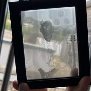 May include: A black framed photograph featuring a portrait of a person in a white suit. The image is a negative, with light areas appearing dark and vice versa. The frame is held by a hand, with a blurred background visible.