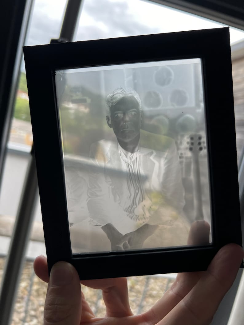 May include: A black framed photograph featuring a portrait of a person in a white suit. The image is a negative, with light areas appearing dark and vice versa. The frame is held by a hand, with a blurred background visible.