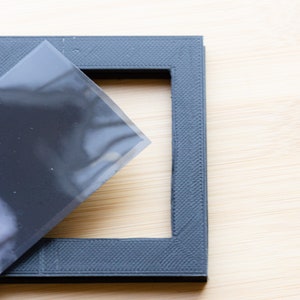 May include: A dark gray picture frame with a textured surface. A sheet of clear plastic partially covers the frame. The frame is square with a rectangular opening.