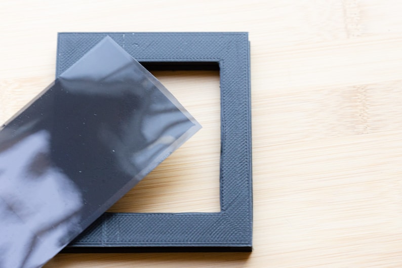 May include: A dark gray picture frame with a textured surface. A sheet of clear plastic partially covers the frame. The frame is square with a rectangular opening.