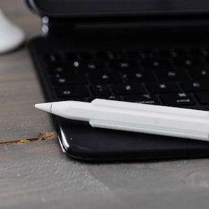 Puede incluir: Un Apple Pencil blanco está sobre un teclado de portátil negro. El portátil está cerrado y el teclado es visible.