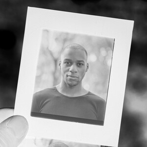 May include: Black and white photograph of a person in a dark shirt. The person is looking at the camera. The photograph is in a white frame, held by a hand. The background is blurred.