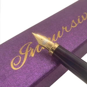 Op de afbeelding: Close-up van een vulpen met een gouden penpunt en een donkerbruine schacht. De pen ligt op een paarse doos met gouden cursieve letters met de tekst "Incursive". De penpunt is scherp.