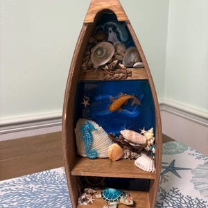 May include: Wooden boat-shaped shelf filled with seashells, a seahorse, a turtle, and a fish against a blue background. The shelf has three levels, creating a miniature ocean scene. The boat is made of light brown wood.