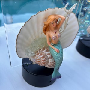 May include: A mermaid figurine sits on a seashell with a light-up base. The mermaid is wearing a green tail and a seashell bra. She has long red hair and is holding a seashell in her hand.