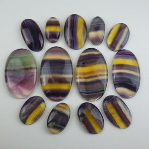 Può includere: Una collezione di 12 pietre di fluorite di forma ovale, in una varietà di colori, tra cui viola, giallo e verde. Le pietre sono lucidate e hanno una finitura liscia e lucida.