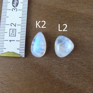 May include: Two polished moonstone cabochons labeled K2 and L2, positioned next to a white measuring tape showing a scale in centimeters. The K2 stone is teardrop-shaped, while the L2 stone is triangular, both displaying a blue iridescent sheen.