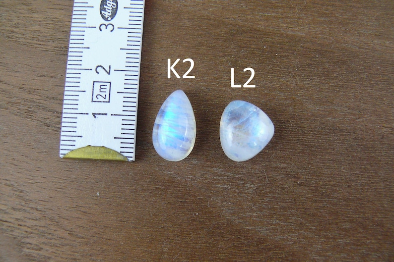 May include: Two polished moonstone cabochons labeled K2 and L2, positioned next to a white measuring tape showing a scale in centimeters. The K2 stone is teardrop-shaped, while the L2 stone is triangular, both displaying a blue iridescent sheen.