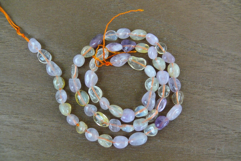 May include: A strand of light pink, yellow, and purple gemstone beads. The beads are smooth and oval-shaped. The beads are strung on a thin orange thread.