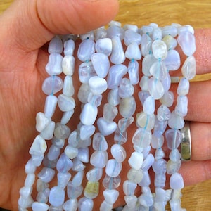 Puede incluir: Múltiples hilos de cuentas de calcedonia azul claro, cada una con una forma suave, pulida e irregular similar a un guijarro. Las piedras azul pálido translúcidas están ensartadas en un hilo fino de color azul claro, creando un aspecto uniforme.