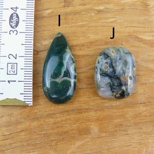 May include: Two polished gemstones labeled I and J next to a ruler. Gemstone I is a dark green teardrop shape with white inclusions. Gemstone J is an oval shape with a mix of grey, green, and black patterns. The ruler shows a scale in centimeters.