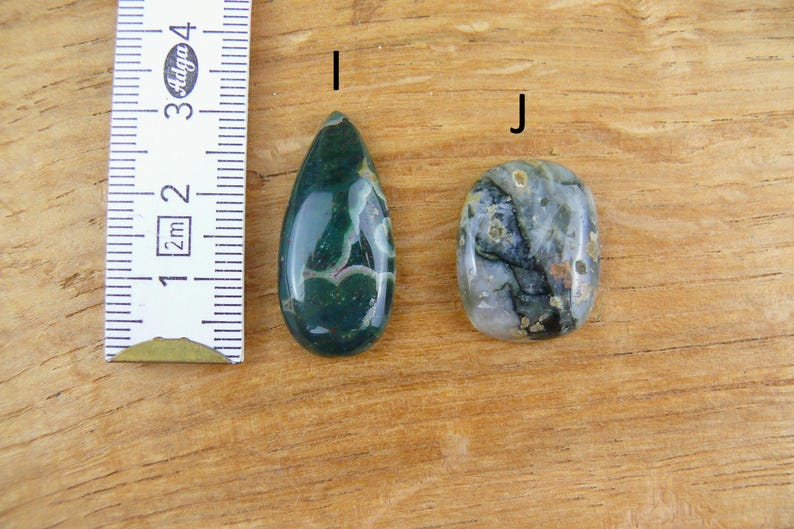 May include: Two polished gemstones labeled I and J next to a ruler. Gemstone I is a dark green teardrop shape with white inclusions. Gemstone J is an oval shape with a mix of grey, green, and black patterns. The ruler shows a scale in centimeters.