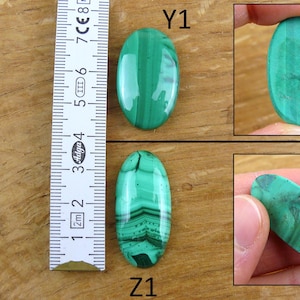 May include: Two oval-shaped malachite gemstones, one with a solid green color and the other with banded patterns. A ruler shows the stones' length. The gemstones are displayed on a wooden surface.