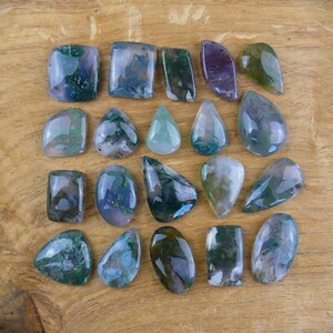 Può includere: Una collezione di pietre preziose lucide di varie forme, tra cui rettangoli, ovali e gocce. Le pietre presentano un mix di colori, con verdi e blu dominanti, e alcuni accenni di viola e bianco. Le pietre preziose sono disposte su una superficie di legno.
