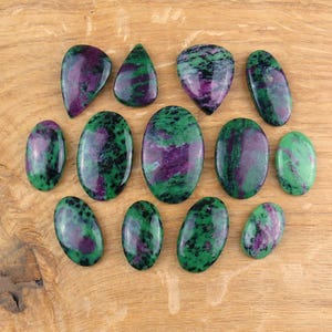Puede incluir: Colección de gemas pulidas de rubí zoisita en varias formas, incluyendo ovaladas y de lágrima, con una mezcla de tonos verdes y morados. Las gemas están dispuestas sobre una superficie de madera marrón claro.