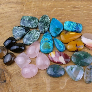 Può includere: Una collezione di pietre preziose levigate di varie forme e dimensioni, tra cui pietre verde scuro, blu brillante, rosa traslucido, giallo, marrone e verde muschio. Queste pietre lisce in stile cabochon presentano motivi naturali distinti.
