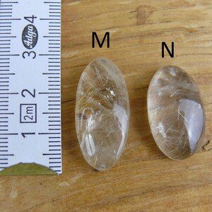 May include: Two oval-shaped clear quartz crystals with gold inclusions, labeled "M" and "N", next to a ruler measuring 3 cm.