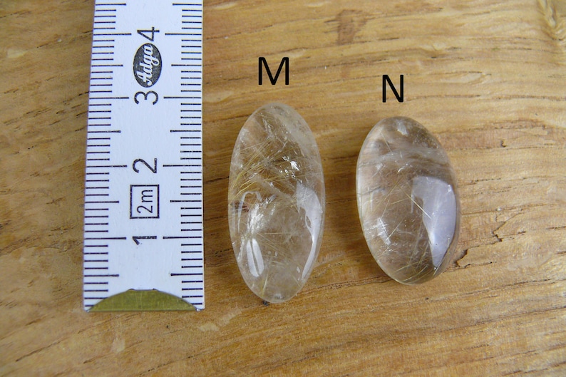 May include: Two oval-shaped clear quartz crystals with gold inclusions, labeled "M" and "N", next to a ruler measuring 3 cm.