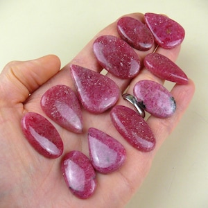 Può includere: Una collezione di undici pietre preziose levigate a forma di goccia e ovale, di colore rosa e magenta, con una finitura liscia e lucida. Le pietre presentano variazioni naturali nell'intensità del colore e nella consistenza, mostrando una tonalità rosa vivace.