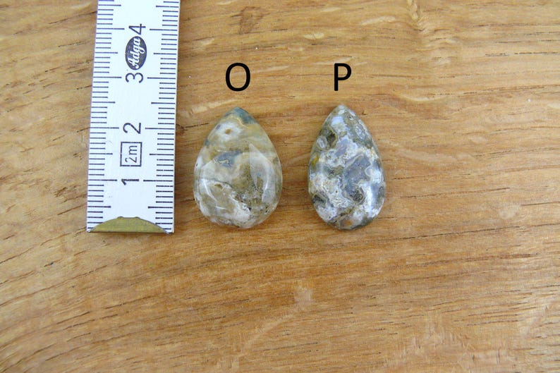 May include: Two teardrop-shaped polished stones labeled O and P, positioned next to a white ruler showing a measurement of approximately 1.5 inches. The stones feature a mottled pattern of gray, beige, and white tones with a smooth, glossy finish.