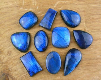 May include: An assortment of polished labradorite gemstones in various shapes, including oval, rectangular, and teardrop. The stones display a vibrant blue iridescence, set against a natural wood grain background. These gemstones are ideal for jewelry making.